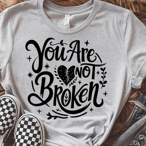 May include: Light gray t-shirt with the words "You Are Not Broken" in a black, decorative font. The design includes a heart and floral elements. Checkered sneakers are visible at the bottom of the image.