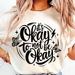 May include: Cream-colored t-shirt with the phrase "It's Okay to Not Be Okay" in a black, circular design. The design includes butterflies, stars, and a lightning bolt. The shirt is a casual wear item.