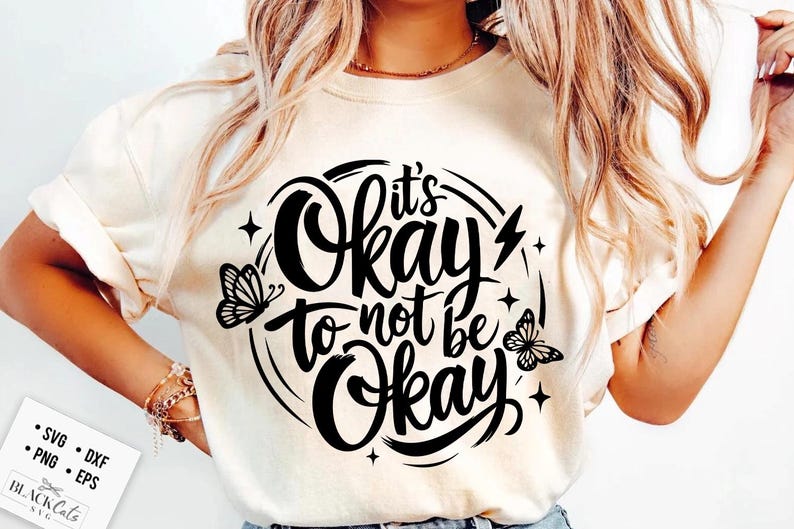 May include: Cream-colored t-shirt with the phrase "It's Okay to Not Be Okay" in a black, circular design. The design includes butterflies, stars, and a lightning bolt. The shirt is a casual wear item.