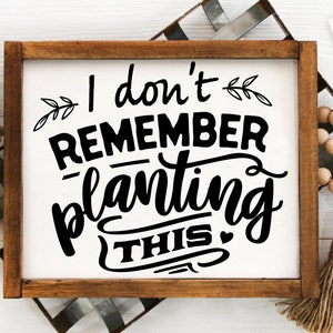 May include: A white wooden sign with a black inscription that reads "I don't remember planting this." The sign is framed in a dark wood frame.