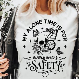 以下が含まれることがあります： 蝶の羽、バラ、そして「MY ALONE TIME IS FOR everyone's SAFETY」の文字が描かれた、白いTシャツ。黒い革のジャケットを着ています。