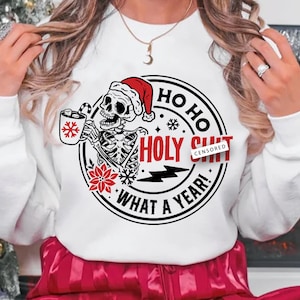 Puede incluir: Sudadera blanca con un diseño circular negro y rojo que presenta un esqueleto sosteniendo una taza con un copo de nieve y el texto "HO HO HOLY SHIT WHAT A YEAR!" con la palabra "SHIT" censurada.