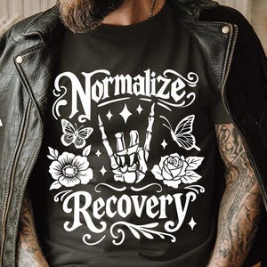 Puede incluir: Camiseta negra con las palabras "Normalize Recovery" en blanco, junto con una mano de esqueleto, mariposas y diseños florales. La persona lleva una chaqueta de cuero negra.