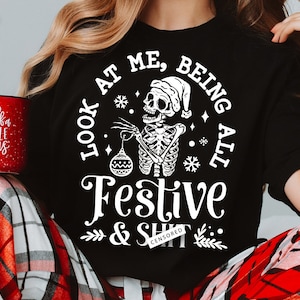 May include: Black t-shirt with a skeleton wearing a Santa hat and holding a bauble. The text reads "LOOK AT ME, BEING ALL Festive & SHIT CENSORED". A red mug with white text is held in the person's hand.