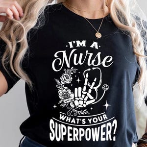 Puede incluir: Camiseta negra con texto blanco que dice "I'm a Nurse What's Your Superpower?" El texto está rodeado de un diseño floral y una mano de esqueleto sosteniendo un estetoscopio.