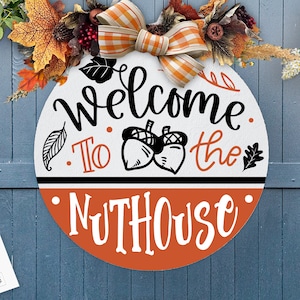 May include: A round wooden sign with a white background and orange and black lettering. The sign says "Welcome to the Nuthouse". There are two acorns and leaves on the sign. The sign is decorated with a fall-themed wreath.