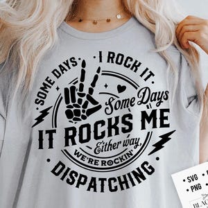 May include: Light gray t-shirt with a black graphic that reads "Some days I rock it, some days it rocks me, either way we're rockin' dispatching." The design includes a skeleton hand making a peace sign. A small white card with "SVG, DXF, PNG, EPS" is in the bottom right.