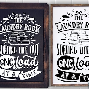 May include: Two black and white signs with the text "The Laundry Room Sorting Life Out One Load At A Time". The signs are on a white tiled background with a green plant in the corner.