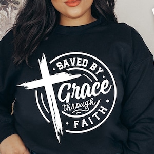 May include: A black sweatshirt with a white graphic design of a cross and a circle with the text "Saved by Grace through Faith".