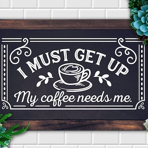 Pode incluir: Um letreiro preto de ardósia com texto branco que diz "I MUST GET UP My coffee needs me." O letreiro é emoldurado em madeira e tem uma borda decorativa. Uma xícara de café com um desenho de coração é representada no centro do letreiro.