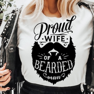 Peut inclure: Un t-shirt blanc avec un texte noir qui dit "Proud Wife of a Bearded Man". Le texte est dans une police de caractères stylisée et est entouré d'un graphique de barbe noire.
