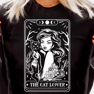 May include: Black long-sleeve shirt featuring a white graphic of a woman holding a black cat, with the text "THE CAT LOVER." The design includes floral and celestial elements. The shirt also has the text "SVG, DXF, PNG, EPS" and the logo "BLACK Cats SVG".