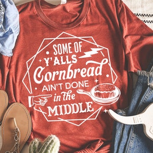Peut inclure: T-shirt rouge avec un motif graphique blanc qui dit "Some of y'alls cornbread ain't done in the middle". Le graphique comprend également un éclair et une poêle avec du pain de maïs à l'intérieur.