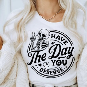 May include: A white t-shirt with a black and white graphic design featuring a skeleton giving the peace sign and the text "Have the Day You Deserve."