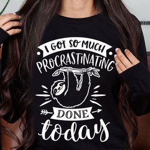 Könnte beinhalten: Schwarzes Sweatshirt mit einer weißen Grafik eines Faultiers, das an einem Ast hängt. Der Text "Ich habe so viel prokrastiniert, dass ich heute fertig bin" steht um das Faultier herum.