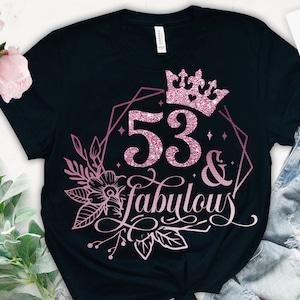 May include: A black t-shirt featuring a pink glitter design that reads "53 & fabulous" with a crown above the number. The design is enclosed within a geometric frame and adorned with floral accents.