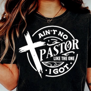May include: A black t-shirt with a white circle design featuring a cross, the text "Ain't No Pastor Like The One I Got." and a stylized flourish.