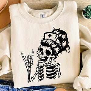 Puede incluir: Sudadera de color crema con un gráfico de enfermera esqueleto en blanco y negro. El esqueleto hace el gesto de la mano de rock and roll y lleva un gorro de enfermera. La sudadera está hecha de un material suave.