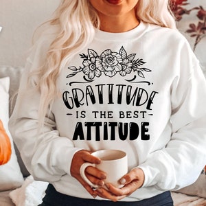 以下が含まれることがあります： 黒い文字で「Gratitude is the best attitude」と書かれた白いスウェットシャツ。テキストの上に花柄のデザインが施されています。