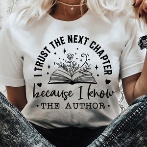 May include: A white t-shirt with a black graphic design featuring an open book with flowers growing from it. The text reads "I trust the next chapter because I know the author."