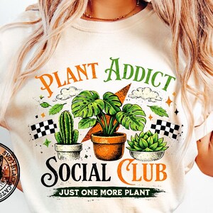 May include: Cream-colored t-shirt with a graphic featuring the text "PLANT ADDICT SOCIAL CLUB" and illustrations of plants, including a cactus, a monstera, and a succulent. The design includes clouds, stars, and checkered flags.