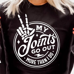 May include: Black sweatshirt with a white graphic that reads "MY Joints GO OUT MORE THAN I DO." The design includes a skeleton hand making a peace sign. The sweatshirt is worn by a person with blonde hair.