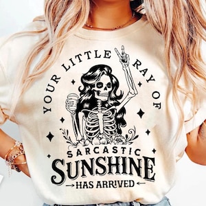 May include: Cream-colored t-shirt featuring a black and white graphic of a skeleton woman holding a coffee cup and giving a peace sign. The text reads "YOUR LITTLE RAY OF SARCASTIC SUNSHINE HAS ARRIVED."