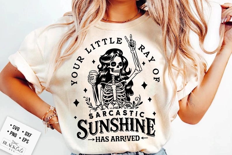 May include: Cream-colored t-shirt featuring a black and white graphic of a skeleton woman holding a coffee cup and giving a peace sign. The text reads "YOUR LITTLE RAY OF SARCASTIC SUNSHINE HAS ARRIVED."