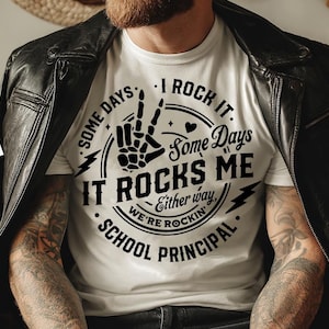 Może przedstawiać: Biała koszulka z napisem "SOME DAYS I ROCK IT. Some Days IT ROCKS ME Either way WE'RE ROCKIN' SCHOOL PRINCIPAL" w czarnym wzorze. Koszulka jest noszona pod czarną skórzaną kurtką.