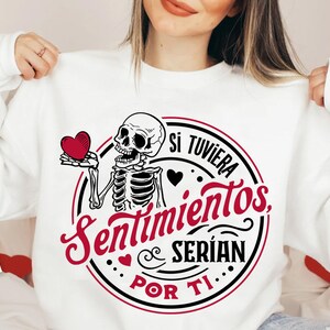 May include: A white sweatshirt with a black and red graphic design featuring a skeleton holding a red heart. The graphic text reads "Si tuviera sentimientos, serian por ti."