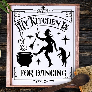 May include: A framed sign with the text "My Kitchen Is For Dancing" in a decorative font. The black and white design features a witch with a broom, a cauldron, and a cat. The sign is on a wooden surface with other decorative items.