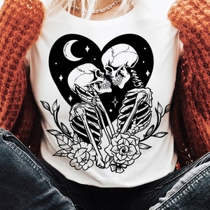 May include: A white t-shirt featuring a black and white graphic of two skeletons kissing within a heart shape. The heart is surrounded by stars and a crescent moon. The skeletons are holding flowers. The text "SVG - DXF - PNG - EPS" is in the bottom right corner of the image.