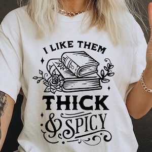 Peut inclure: Un t-shirt blanc avec du texte noir qui dit "J'aime qu'ils soient épais et épicés" avec une pile de livres et un motif floral.