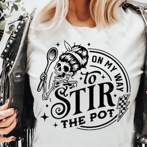 May include: White t-shirt with a black graphic of a skeleton holding a spoon, with the text "ON MY WAY to STIR THE POT". The shirt is worn with a black leather jacket.