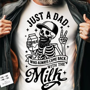 May include: White t-shirt featuring a black graphic of a skeleton wearing a cap and holding a milk carton, with the text "JUST A DAD WHO ALWAYS CAME BACK WITH THE MILK". The design includes stars and a banner.