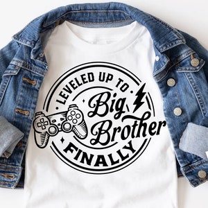 May include: A white t-shirt with a black and white graphic design of a video game controller inside a circle with the text "Leveled Up To Big Brother Finally".