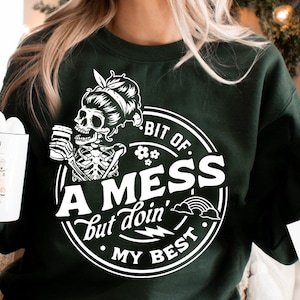 May include: Dark green sweatshirt with a white graphic featuring a skeleton drinking from a mug, with the text "A bit of a mess but doin' my best." A mug with marshmallows and the text "Snow Much Fun" is held in the left hand.