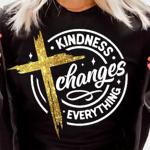 May include: A black t-shirt with a white circle design featuring a gold glitter cross and the text "Kindness changes everything."