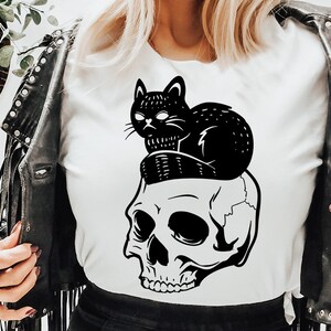 May include: Black and white graphic design featuring a skull with a black cat sitting on top of it. The cat is looking to the left. The design is on a white t-shirt.