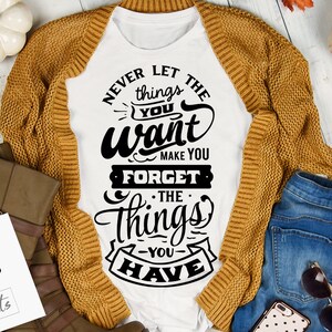 以下が含まれることがあります： 白いTシャツに、「欲しいものが、持っているものを忘れさせることはない」という黒いグラフィックがプリントされています。Tシャツは、木製の表面に、茶色のカーディガン、青いジーンズ、秋の装飾品と一緒に置かれています。