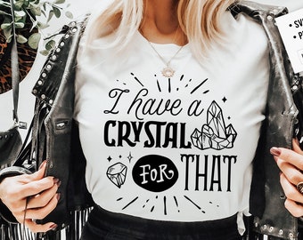 May include: White t-shirt with the phrase "I have a CRYSTAL FOR THAT" in black. The design includes crystal illustrations and starburst accents. The shirt is worn with a black leather jacket.