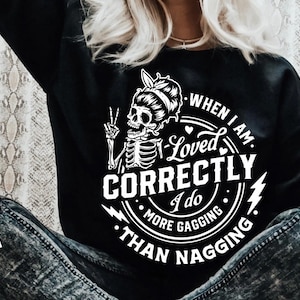 May include: Black sweatshirt with a white skeleton graphic giving a peace sign and the text "When I am loved correctly I do more gagging than nagging." A Black Cats SVG logo is in the lower left corner.