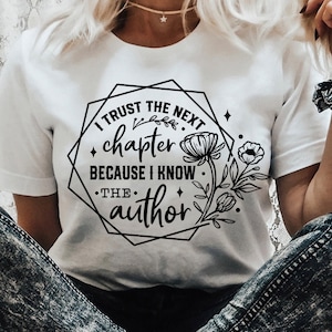 May include: White t-shirt with black text that reads "I trust the next chapter because I know the author." The text is surrounded by a geometric shape with floral accents.