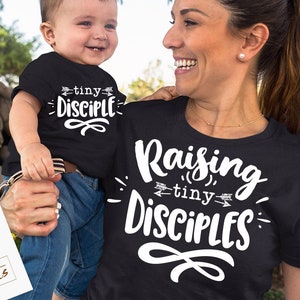 May include: A mother and child wearing black t-shirts with white text. The child's shirt says "tiny disciple" and the mother's shirt says "Raising tiny disciples".