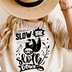 May include: A white t-shirt with a black graphic of a sloth hanging from a branch with the text "Slow the sloth down".
