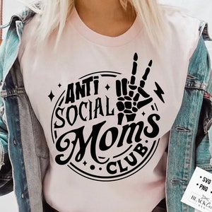Op de afbeelding: Een roze T-shirt met een zwart cirkelvormig ontwerp met de tekst "Anti Social Moms Club" en een skelet hand die een peace-teken maakt.