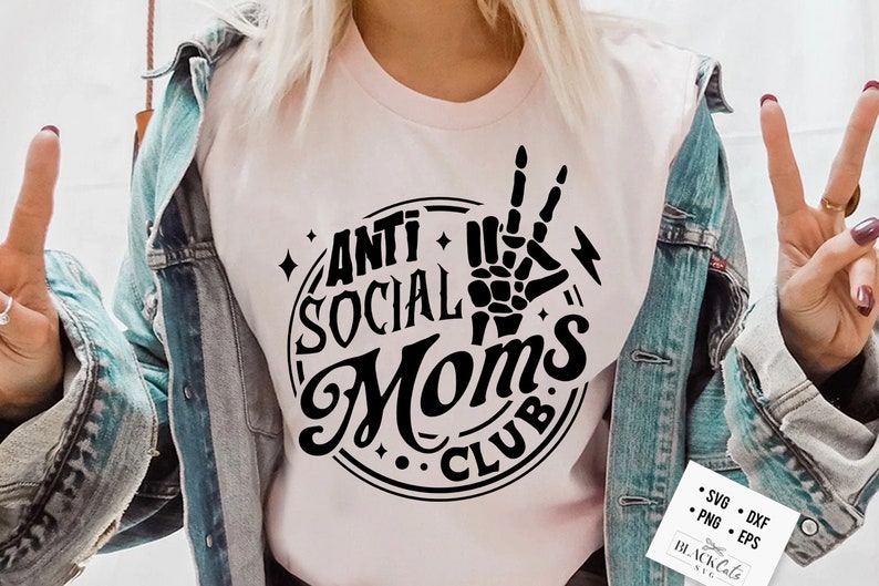 Op de afbeelding: Een roze T-shirt met een zwart cirkelvormig ontwerp met de tekst "Anti Social Moms Club" en een skelet hand die een peace-teken maakt.
