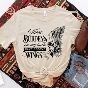 Op de afbeelding: Een beige T-shirt met een zwart grafisch ontwerp met een engel met vleugels en de tekst "These Burdens on my back Have Become Wings".