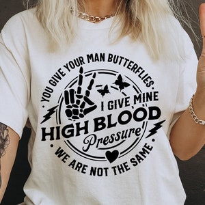 May include: A white t-shirt with a black graphic design. The graphic features a peace sign with a skeleton hand, a lightning bolt, butterflies, and the text "You give your man butterflies. I give mine high blood pressure. We are not the same."