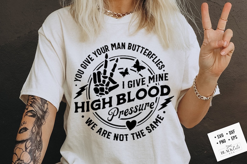 May include: A white t-shirt with a black graphic design. The graphic features a peace sign with a skeleton hand, a lightning bolt, butterflies, and the text "You give your man butterflies. I give mine high blood pressure. We are not the same."
